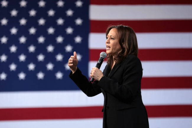 Kamala Harris speaks into a microphone while gesturing toward an unseen audience in front of an American flag.