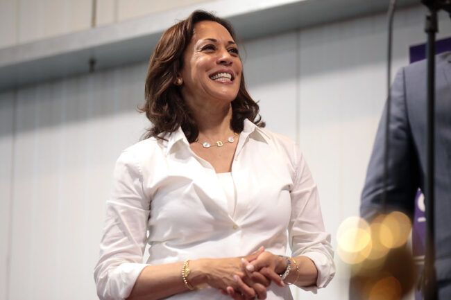 Kamala Harris smiles while wearing a white collared shirt and necklace, looking to the upper right toward an unseen person.