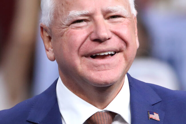 Governor Tim Walz smiles wearing a blue suit, white shirt, and American flag label pin.